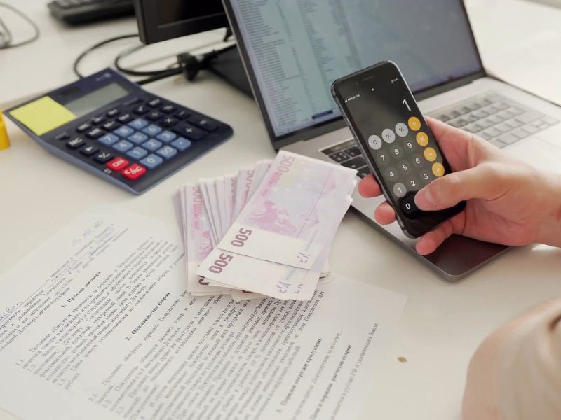osoba sedi na stolu na kom je laptop, dokumentacija za kredit i velika količina novca, računa na telefonu nešto